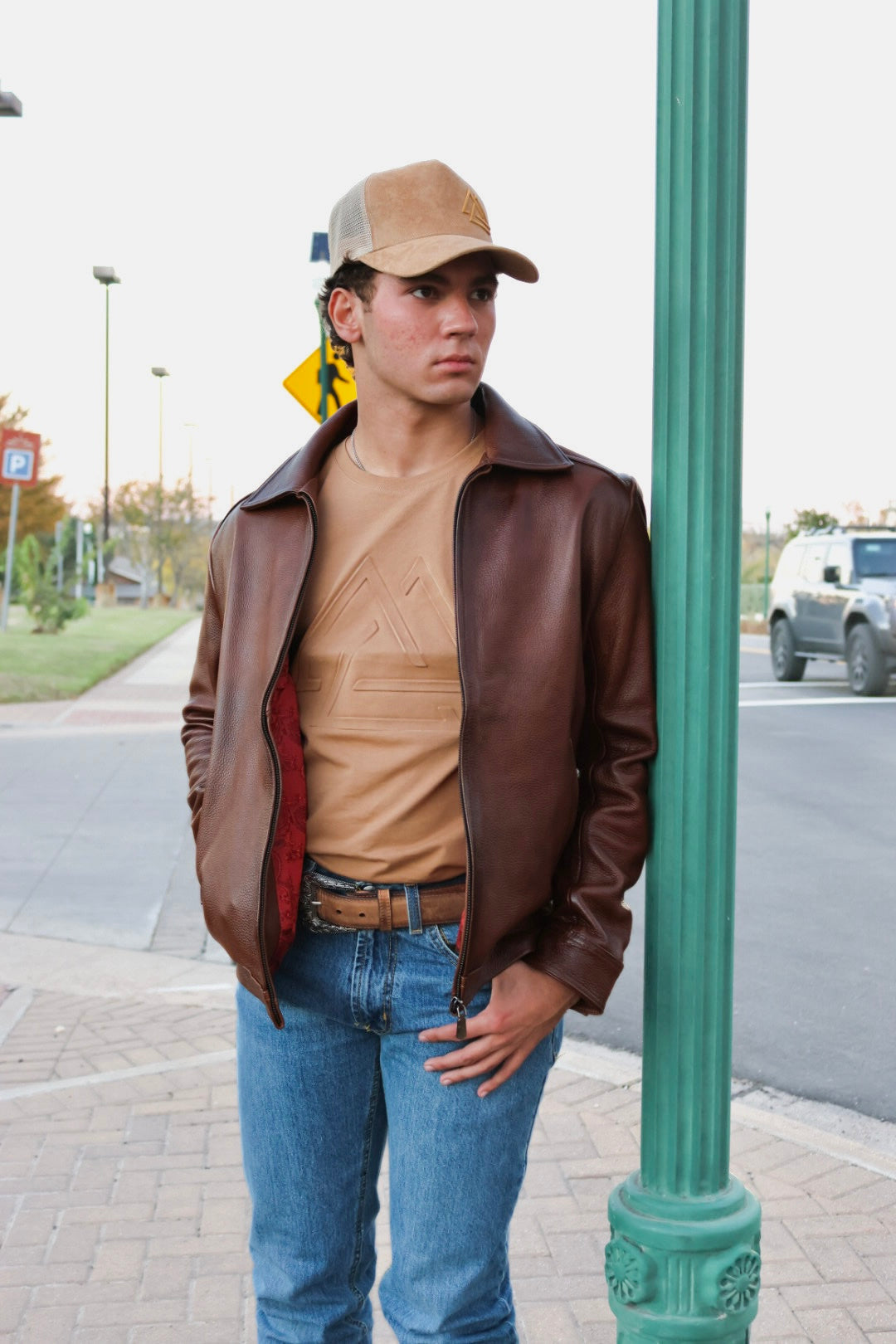Person wearing a brown leather jacket, beige cap, leather belt and blue jeans