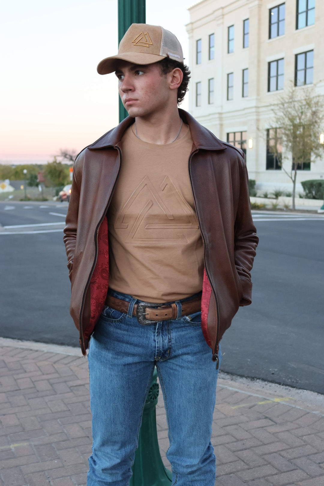 Person wearing a brown leather jacket, beige shirt, blue jeans, and a leather belt on a street.