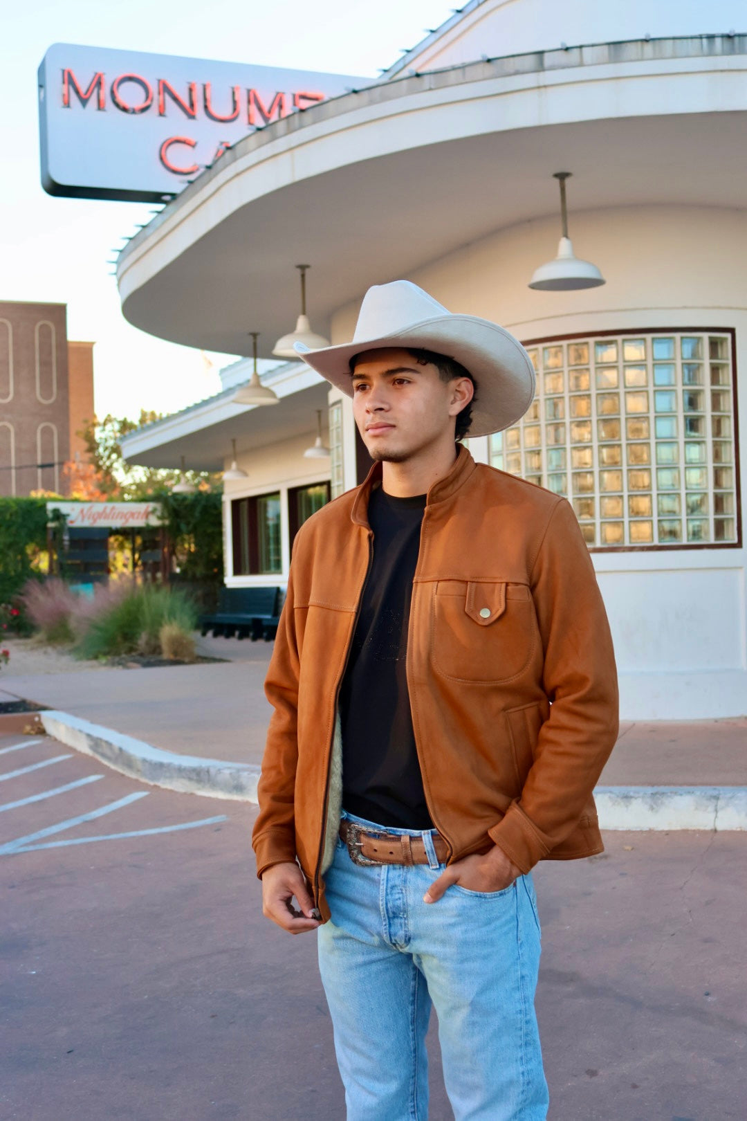 Man wearing a cowboy hat and brown jacket in front of a building with 'Monum' sign.
